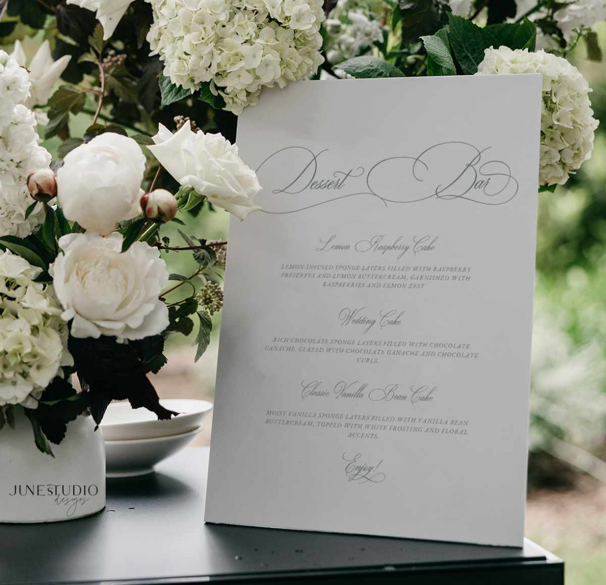 A white wedding invitation card with elegant gray script text stands on a dark tabletop, surrounded by white flowers and green foliage in a real outdoor setting.