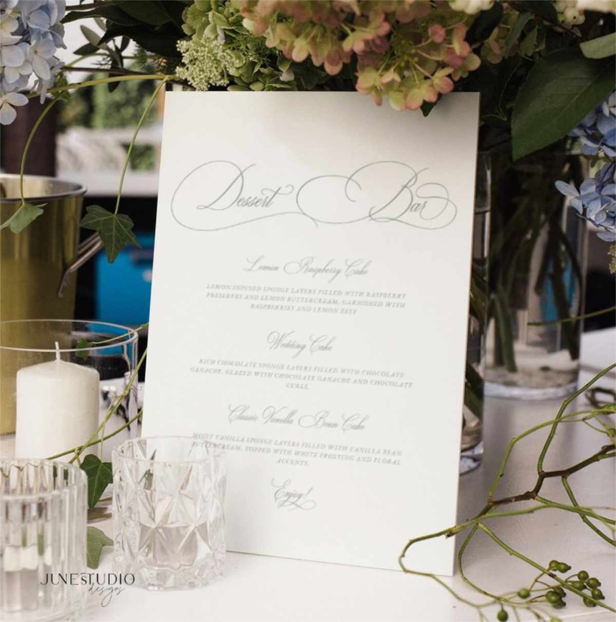 A white wedding reception program or menu card with elegant gray script lettering, styled on a table with glass candleholders, flowers, and greenery.