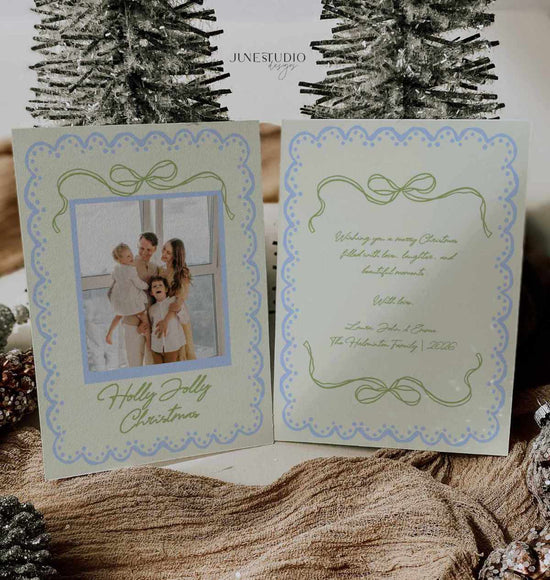 A double-sided Christmas greeting card set displayed open, showing a photo of a family on the front and elegant script text on the back. The card has a pale green and blue scalloped border with bow motifs.