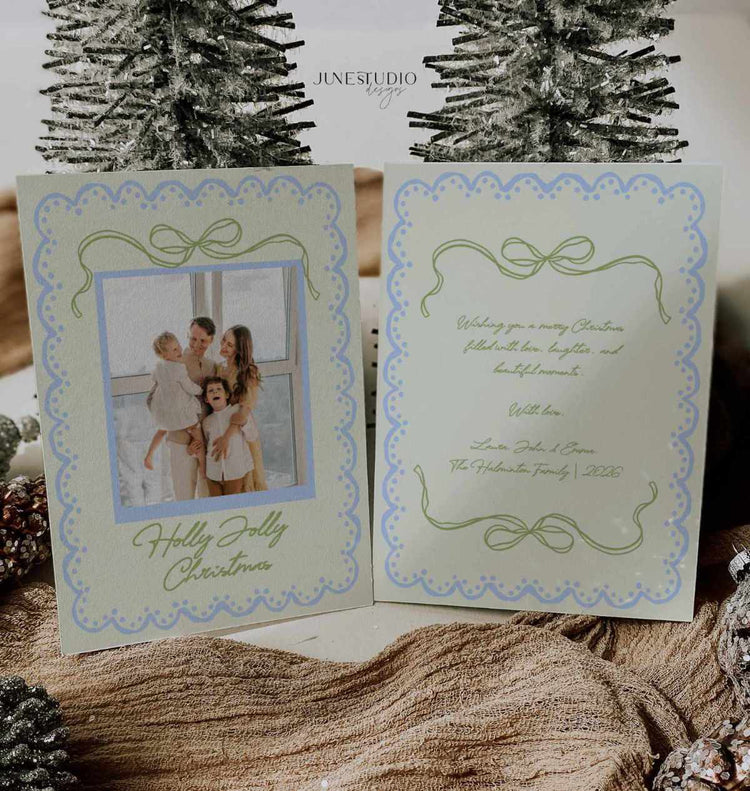 A double-sided Christmas greeting card set displayed open, showing a photo of a family on the front and elegant script text on the back. The card has a pale green and blue scalloped border with bow motifs.