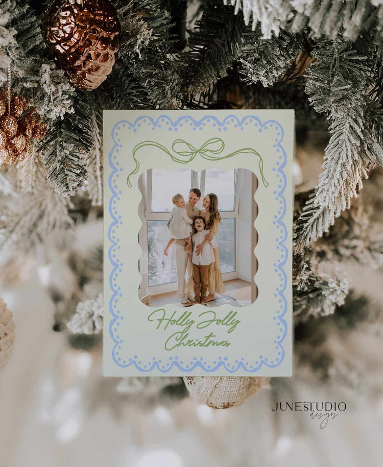 A personalized holiday photo card with a scalloped blue border and green script text reading 'Holly Jolly Christmas,' hanging on a decorated Christmas tree with pinecones and frosted branches.